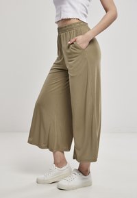 Personne portant un pantalon large court kaki avec taille élastique et des baskets blanches, main dans la poche, debout sur un sol blanc.