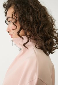 Helles Rosa, Sweatshirt mit hohem Kragen und Reißverschlussdetail. Der Stoff wirkt weich und hat eine gerippte Textur am Kragen. Lockige Haare teilweise sichtbar.