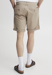 Beige Baumwollshorts mit umgeschlagenen Säumen, ausgestattet mit zwei Gesäßtaschen und Knopfdetails, kombiniert mit weißen Socken und braunen Sandalen.