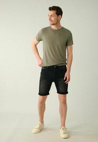 T-shirt ajusté vert olive associé à un short en denim foncé avec un ourlet roulé. Le modèle porte des baskets beiges. L'arrière-plan neutre met en valeur les détails des vêtements.