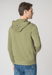 Felpa verde oliva con una vestibilità aderente, maniche lunghe e cappuccio con coulisse. Presenta un piccolo logo sul retro. Indossata con jeans blu.
