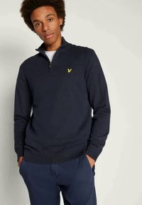 Jūros mėlynumo megztinis su ketvirčio užtrauktuku. Yra mažas geltonas logotipas kairėje krūtinės pusėje ir pylimo apykaklės bei rankovių. Minkšta textura.