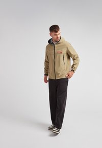 Chaqueta impermeable beige con capucha, cremallera frontal, acentos negros y un logo en el pecho. Combinada con pantalones negros y zapatillas.