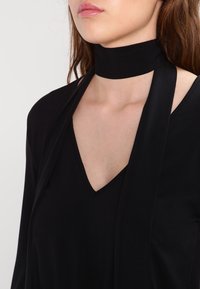 Czarna bluzka z dekoltem w szpic i teksturowanym kołnierzykiem choker. Materiał wydaje się gładki, a projekt zawiera długie, zwisające paski przy szyi.