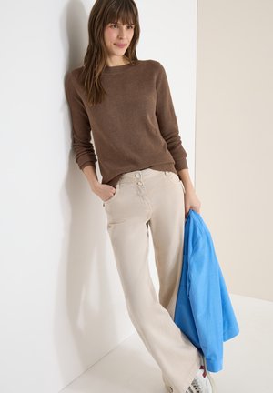 Donna appoggiata a una parete bianca, indossa un maglione marrone, pantaloni beige, sneakers bianche e tiene una giacca blu brillante nella mano destra.