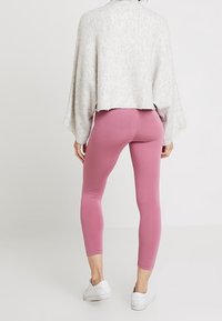 Person som bär en ljusgrå lös tröja, rosa tajta leggings och vita sneakers, står vänd bort från kameran mot en neutral bakgrund.
