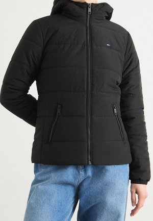 Osoba ubrana w czarną pikowaną kurtkę z przednim zamkiem błyskawicznym i zamykanymi na zamek kieszeniami, w połączeniu z jasnoniebieskimi dżinsami.