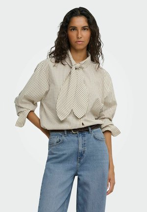 Femme aux longs cheveux bouclés porte un chemisier beige à carreaux avec un grand nœud noué et un jean bleu taille haute sur un fond uni.