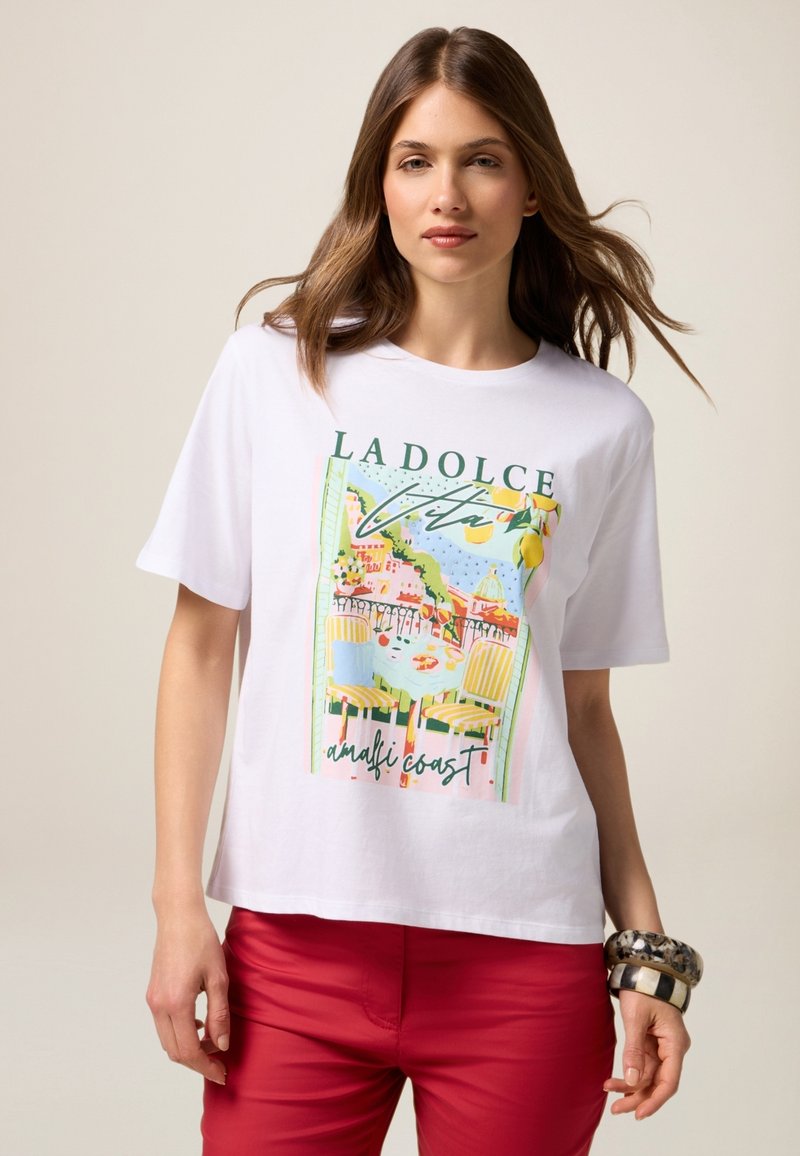 Witte t-shirt met korte mouwen, voorzien van een kleurrijk grafisch ontwerp van de Amalfikust, met een ontspannen pasvorm en een zachte katoenen textuur. In combinatie met rode broek.