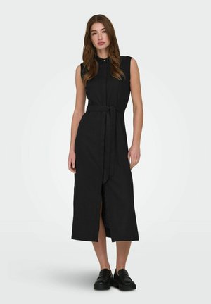 Fiatal nő ujjatlan fekete midi ruhában, elöl hasítékkal és megkötős derékövre, fekete mokaszinokkal párosítva, fehér háttér előtt állva.