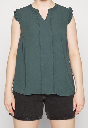 Vrouw die een mouwloze teal blouse met gerimpelde schouders en zwarte denim korte broek draagt, gefotografeerd van de nek tot halverwege de dij tegen een effen achtergrond.