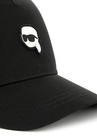 Melnā cepure ar izliekto malu, ar baltu un melnu logotipu ar seju saulesbrillēs priekšpusē. Izgatavota no gludas auduma.
