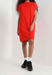 Robe t-shirt en coton rouge avec des manches courtes et un ourlet asymétrique. Portée avec des baskets noires et des chaussettes blanches chevilles.