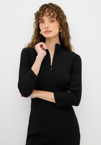 Schwarzes Reißverschlusskleid mit 3/4-Ärmeln, aus glattem Stoff gefertigt. Besitzt einen hohen Kragen und ein schlichtes, figurnahes Design ohne Muster oder Verzierungen.