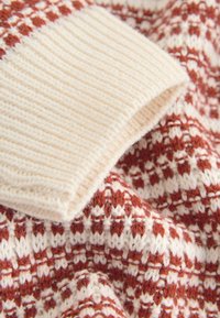 Knitted sweater detail featuring a textured cream cuff and a warm rust-red patterned body. Soft yarn with visible fibers and intricate stitching.