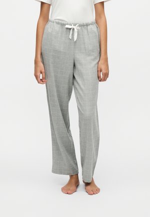 Personne portant un pantalon gris à jambes larges avec un cordon blanc et un motif discret à carreaux, associé à une chemise blanche et pieds nus.