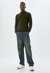 Groene longsleeve trui met een hoge kraag, gecombineerd met vervaagde blauwe jeans. Het model draagt witte sneakers met tan accenten. Eenvoudig, casual ontwerp.