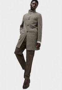 Grå uldfrakke med høj krave og knaplukning, kombineret med brune bukser og mørke støvler. Glat tekstur og skræddersyet pasform.