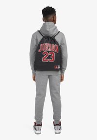 Fekete húzózsinóros hátizsák, rajta piros "JORDAN" és "23" felirattal. Szürke kapucnis pulcsival és melegítőnadrággal viselve, piros részletekkel ellátott sportcipővel kiegészítve.