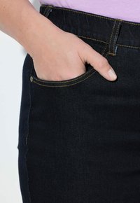 Tmavě modré džíny s viditelným šitím, mají boční kapsu a ruku umístěnou uvnitř. Hladká textura a těsný střih.