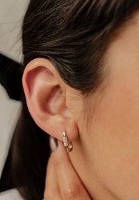 Pendiente de aro dorado con un diseño texturizado y un centro de cristal claro, llevado en el lóbulo de la oreja. Cabello parcialmente visible.