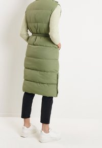 Zelená péřová vesta s páskem v pase, s prošívanými částmi, bočními rozparky a vysokým límcem, v kombinaci s černými kalhotami a bílými teniskami.