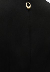 Μαύρο ένδυμα με κεντρική ραφή και ένα μικρό χρυσό οβάλ μενταγιόν προσαρτημένο κοντά στην κορυφή με μαύρο κορδόνι ή αλυσίδα.