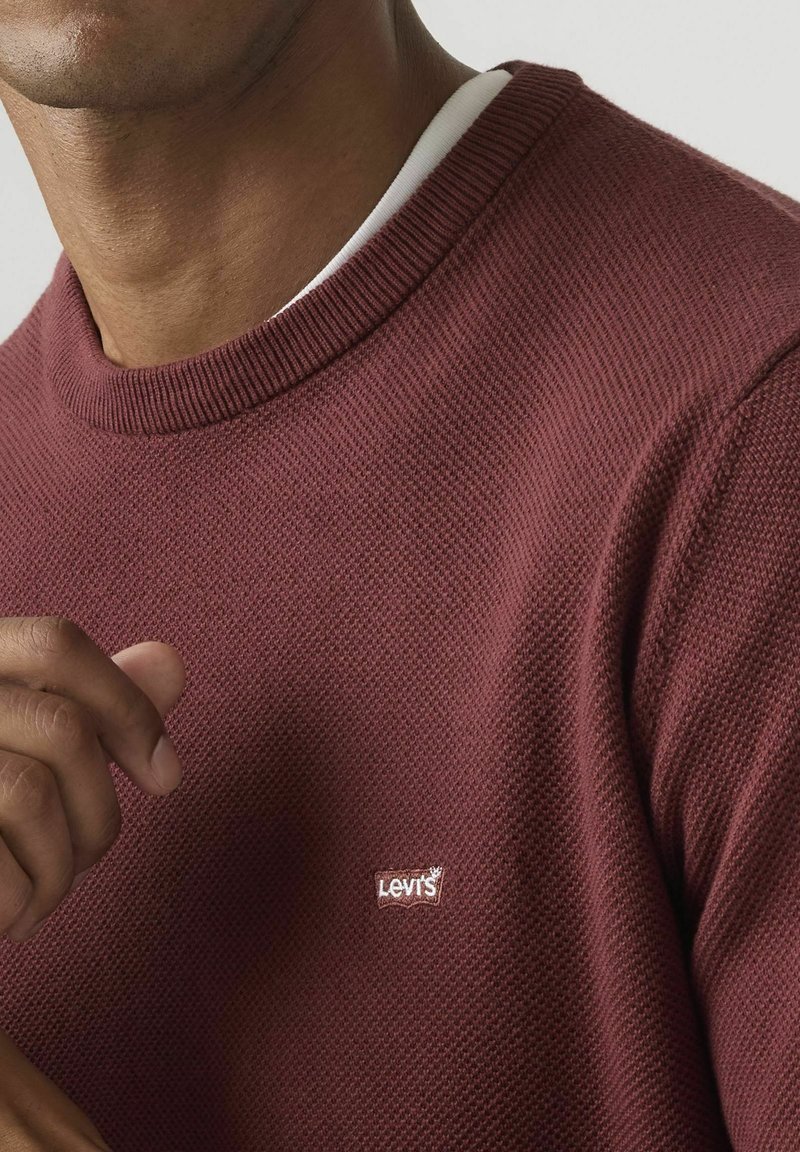 Hombre con un suéter granate texturizado de cuello redondo sobre una camisa blanca, con un pequeño logo blanco de Levi's en el pecho.