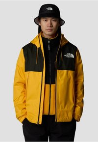 Gelbe und schwarze Jacke mit durchgehendem Reißverschluss, verstellbarem Kapuze und Fleece-Innenfutter; kombiniert mit einem schwarzen Bucket Hat.