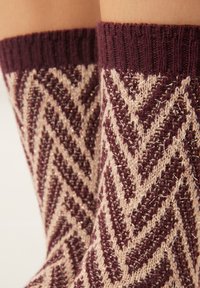 Chaussettes en tricot bordeaux et beige avec un motif en chevrons portées aux pieds sur un fond uni.