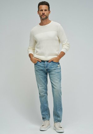 Hombre de pie con las manos en los bolsillos, vistiendo un suéter blanco de punto, jeans desgastados de color azul claro y zapatillas beige sobre un fondo liso.