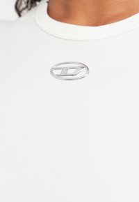 Balta pieguļoša T-krekls ar gludu tekstūru, kakla augšdaļā sudraba metāliska logotipa akcents. Vienkāršs dizains, bez rakstiem.