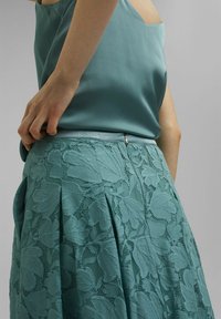 Una blusa verde azulado hecha de una tela suave, combinada con una falda de encaje plisada que presenta patrones de hojas y una cremallera visible en la cintura.