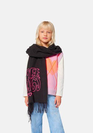 Enfant blond portant un gilet argyle rose, une chemise blanche à manches longues, un jean bleu et une grande écharpe noire avec un texte rose et des franges.