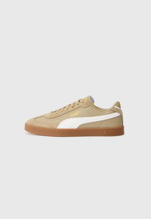 Beige Puma sneaker med hvid stribe, gummisål og gyldent Puma-logo på siden, vist i profil mod en ensfarvet baggrund.