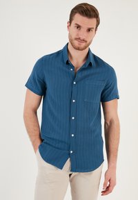 Camisa de manga corta con botones de color azul profundo, hecha de tejido texturizado con rayas verticales, que cuenta con un bolsillo en el pecho y botones blancos.