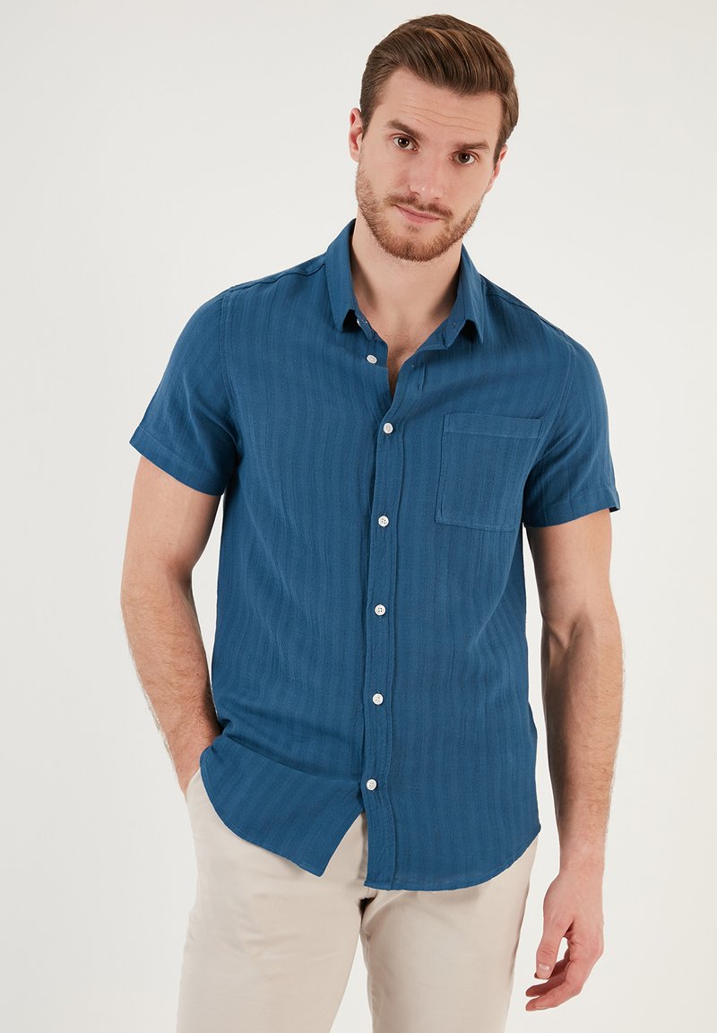 Camisa de manga corta con botones de color azul profundo, hecha de tejido texturizado con rayas verticales, que cuenta con un bolsillo en el pecho y botones blancos.