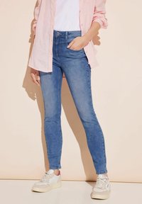 Jean ajustados en denim azul medio con una textura suave, combinados con una camiseta blanca y una camisa de botones rosa claro. Zapatillas blancas casuales.