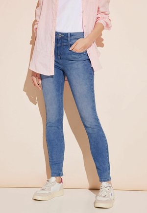 Schmale Jeans aus mittelblauem Denim mit einer glatten Textur, kombiniert mit einem weißen T-Shirt und einem hellrosa Hemd mit Knöpfen. Legere weiße Sneakers.