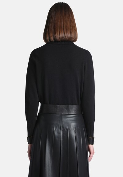 Femme aux cheveux bruns mi-longs portant un top noir à manches longues rentré dans une jupe en cuir noire plissée taille haute, vue de dos.