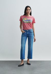Red cotton t-shirt featuring a multicolored graphic print, paired with light blue jeans, and black flats. Simple design, relaxed fit.