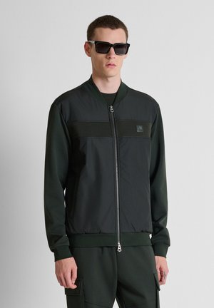 Young man wearing black zippered jacket, dark green pants with pockets, and large black sunglasses, standing against a plain light background.