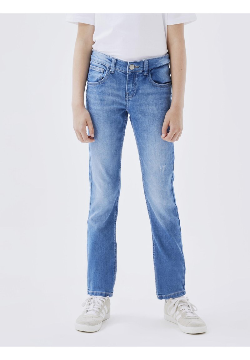 Personne portant un T-shirt blanc uni, un jean droit bleu clair et des baskets blanches, debout devant un fond clair uni.