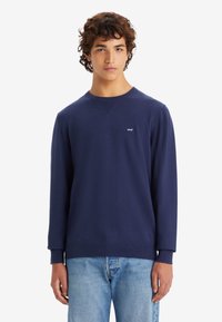 Marineblauwe sweatshirt met lange mouwen, ronde hals, geribbelde accenten en een klein wit logo op de borst. Gecombiniert met lichtblauwe jeans.