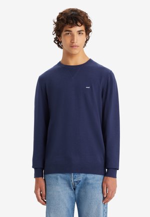 Navy blue long-sleeve sweatshirt with round neck, ribbed accents, and small white logo on chest. Paired with light blue jeans.