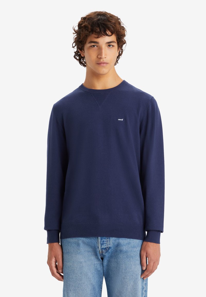 Marineblauwe sweatshirt met lange mouwen, ronde hals, geribbelde accenten en een klein wit logo op de borst. Gecombiniert met lichtblauwe jeans.