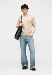 Νεαρός άνδρας που φοράει μπεζ πουλόβερ με κεντημένο πατς αλεπούς, ανοιχτόχρωμα τζιν, μαύρα παπούτσια και κρατάει μαύρη τσάντα ώμου σε λευκό φόντο.