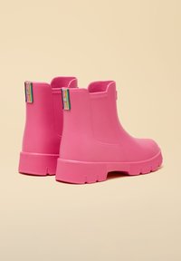 Par lyserøde gummistøvler til anklen med tykke såler og multifarvede stofstropper bagpå, placeret mod en beige baggrund.