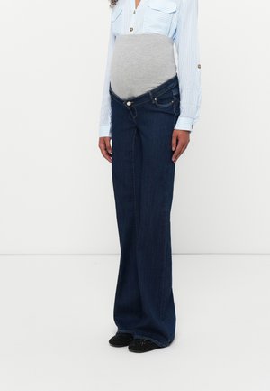Personne enceinte portant une chemise à rayures bleues, une bande de maternité grise, et un jean large bleu foncé déboutonné avec des chaussures noires.