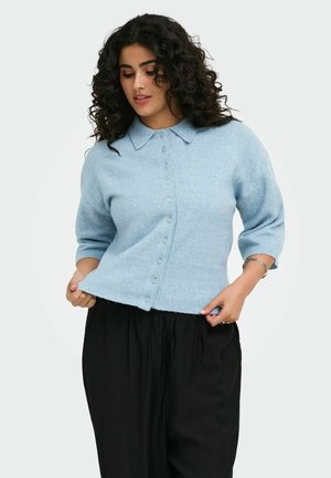 Frau mit lockigem dunklem Haar, die einen hellblauen, geknöpften Pullover mit Kragen und schwarze Hose trägt, passt den Pullover-Saum an.